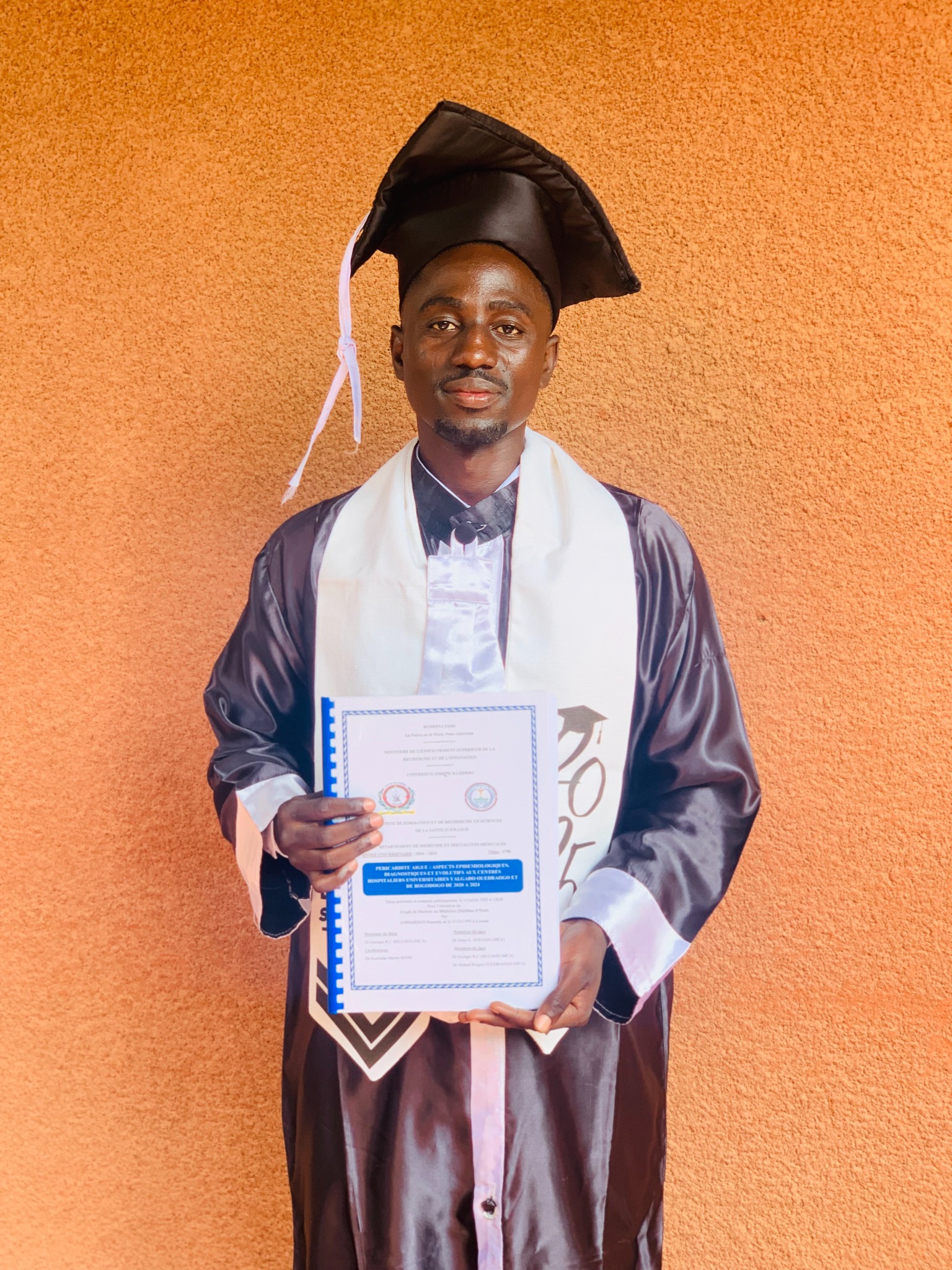 Ousseni Sawadogo en tenue de remise de diplôme, tenant sa thèse de médecine